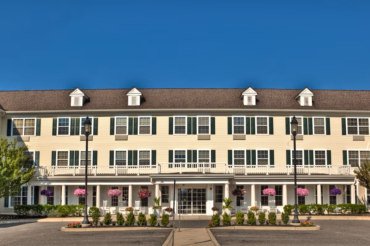 Front exterior view of a three-story senior living facility building with beige siding, green shutters, and a brown roof. The entrance is covered by a porch with white columns and decorated with hanging flower baskets and rocking chairs. Two black street lamps flank the walkway leading to the entrance. The sky is clear and blue.