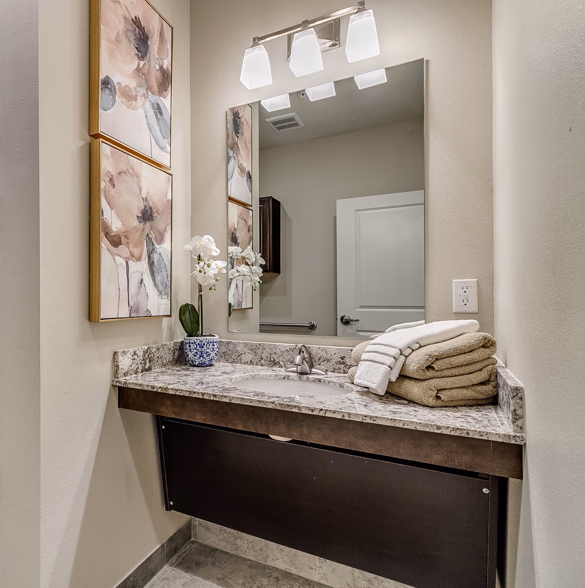 A bathroom vanity with a granite countertop, an under-mount sink, and a silver faucet. On the countertop, there is a small blue and white flower pot with white orchids and a neatly folded stack of beige and white towels. Above the vanity is a large rectangular mirror with a three-light fixture mounted on the wall. Two framed abstract floral paintings hang on the adjacent wall. The bathroom has light-colored walls and a tiled floor.