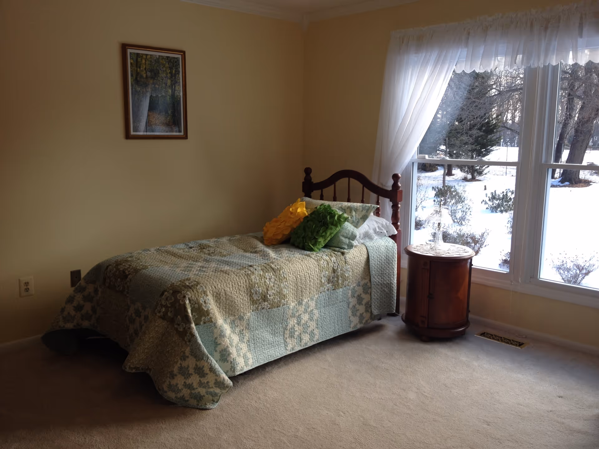 A cozy bedroom with a single wooden bed covered with a patchwork quilt in shades of green and blue, adorned with three decorative pillows in yellow, green, and patterned fabric. Next to the bed is a round wooden nightstand with a lace doily and a glass lamp. A large window with sheer white curtains reveals a snowy outdoor scene with trees and bushes. A framed picture hangs on the beige wall above the bed.