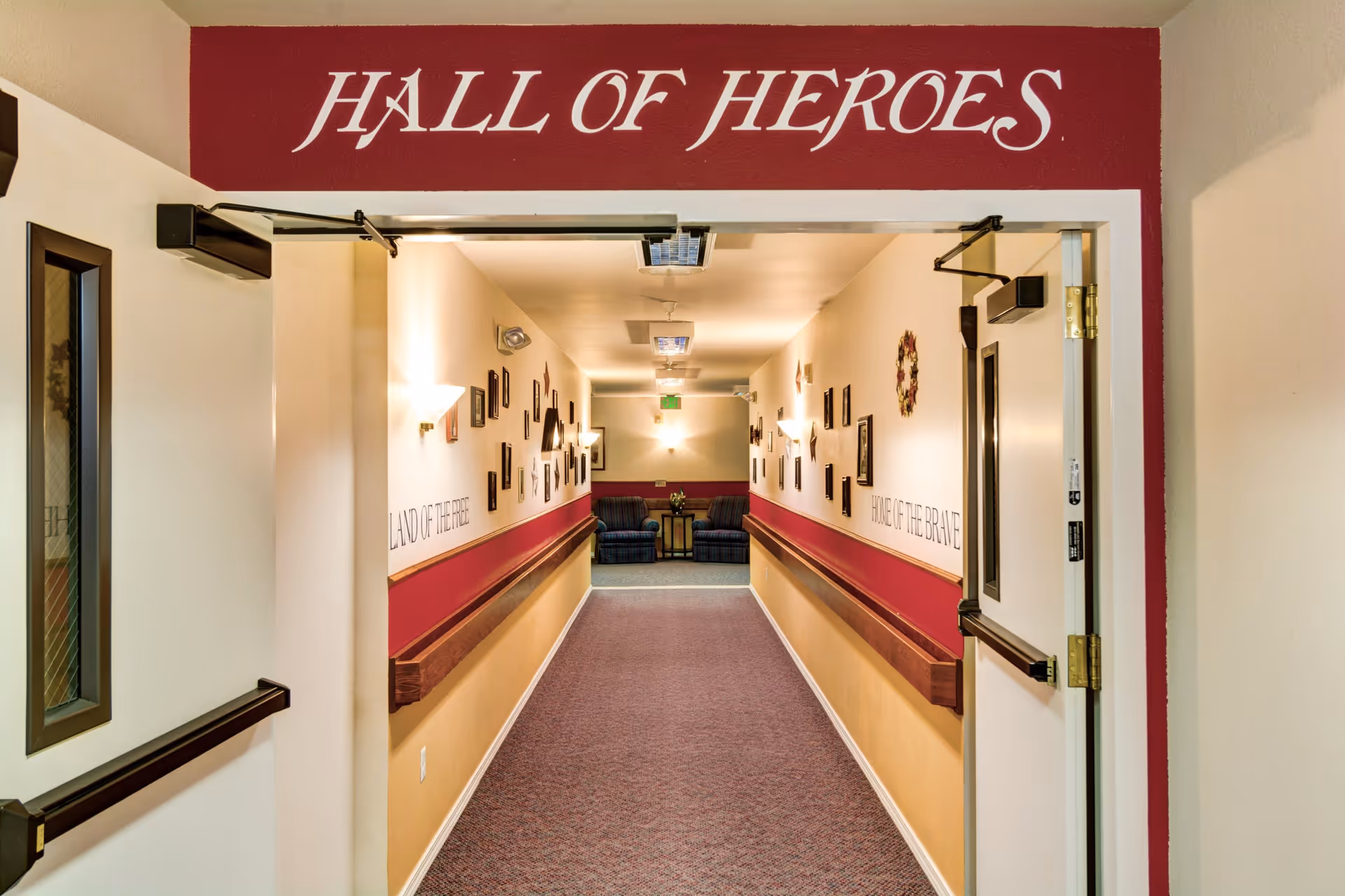 A hallway in a senior living facility with a sign above the entrance reading 'HALL OF HEROES'. The hallway has handrails on both sides and walls decorated with framed pictures and plaques. At the end of the hallway, there are two blue armchairs and a small table with a flower arrangement. The walls have text that reads 'LAND OF THE FREE' on the left and 'HOME OF THE BRAVE' on the right.