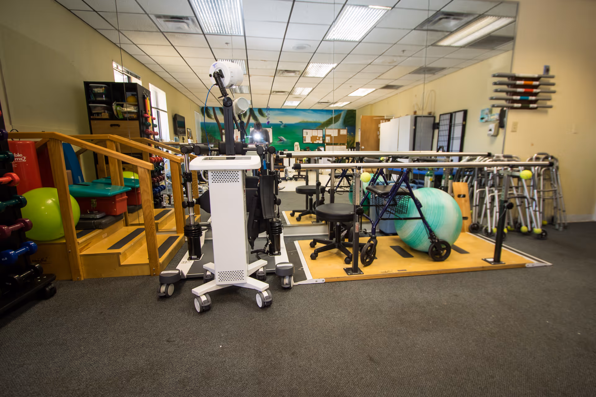 Interior view of a physical therapy or rehabilitation room with exercise equipment including parallel bars, walkers, a large exercise ball, weights, and a small set of stairs with handrails. The room has a carpeted floor, mirrored wall, and fluorescent ceiling lights.