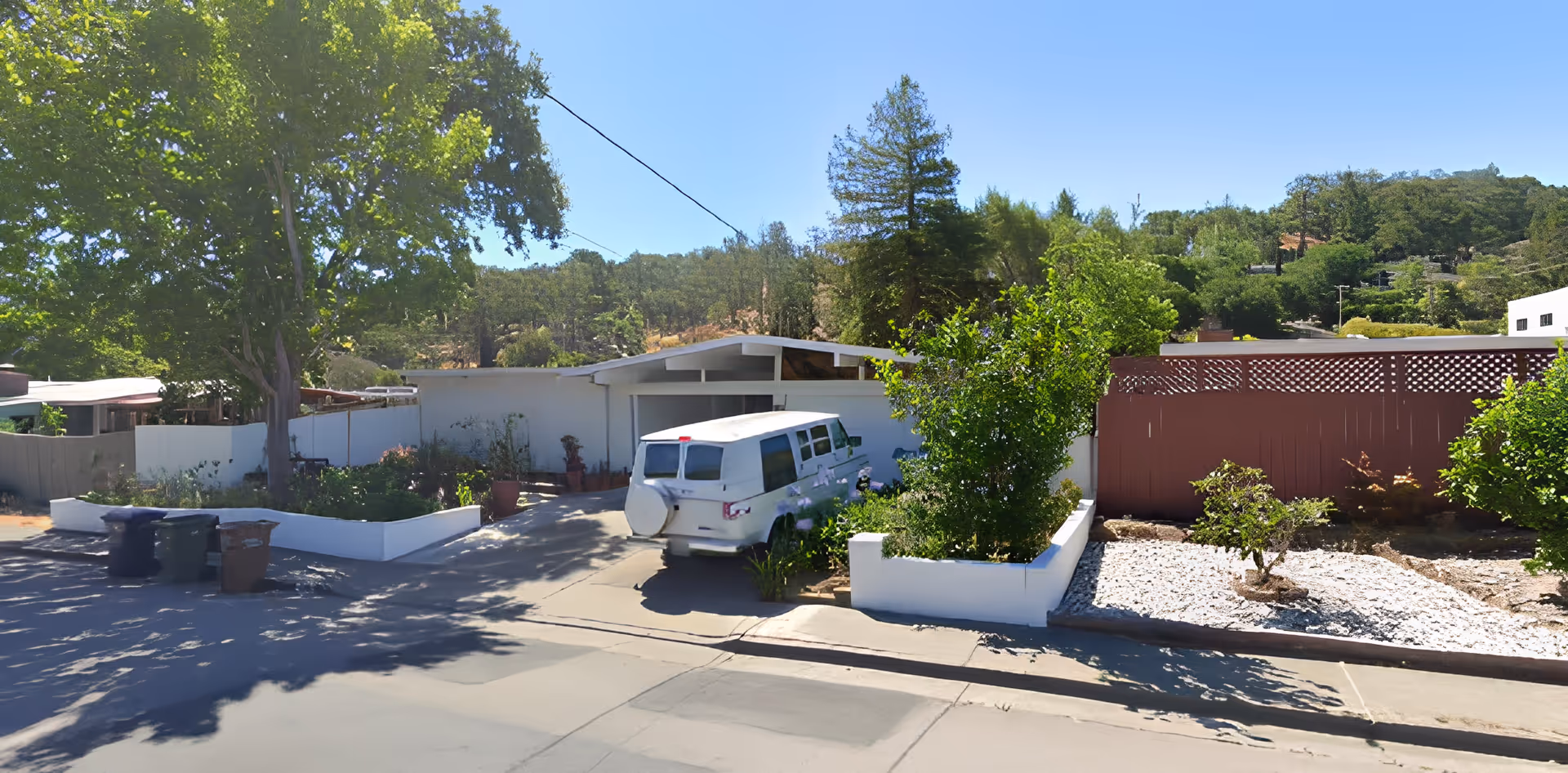 Exterior view of a single-story white building with a driveway and a white van parked in front. The property is surrounded by greenery including trees and bushes, with a red wooden fence on the right side and a clear blue sky above.
