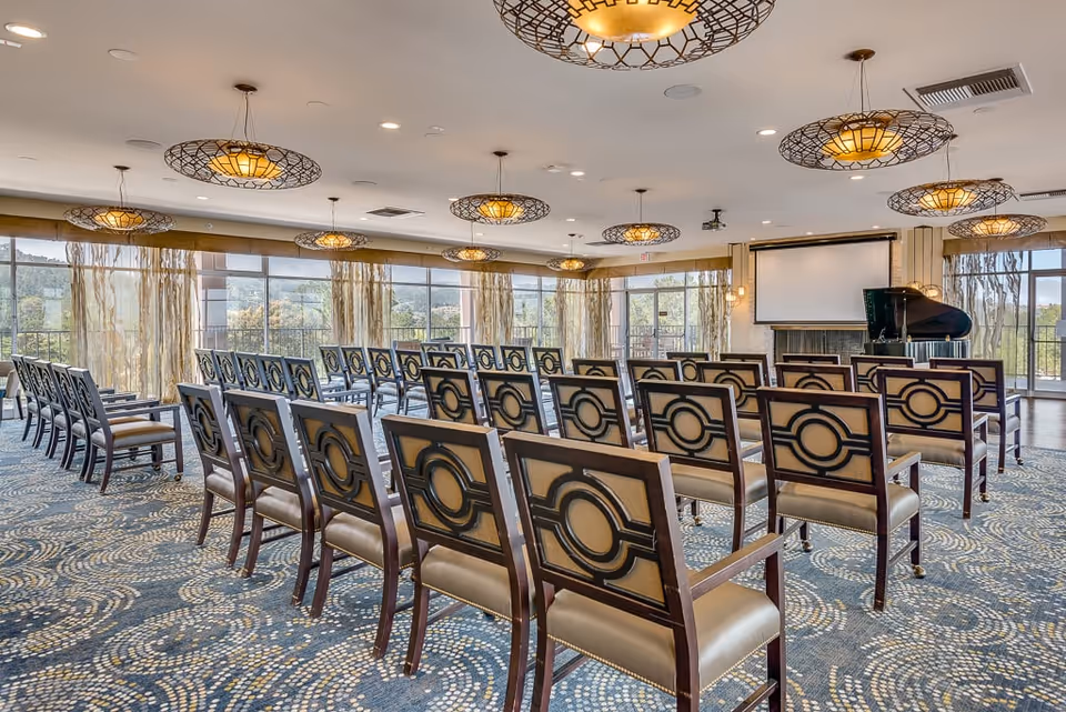 A spacious meeting or event room with rows of wooden chairs featuring circular back designs, a patterned carpet, large windows with sheer curtains allowing natural light, multiple decorative ceiling lights, a grand piano, and a projector screen at the front.
