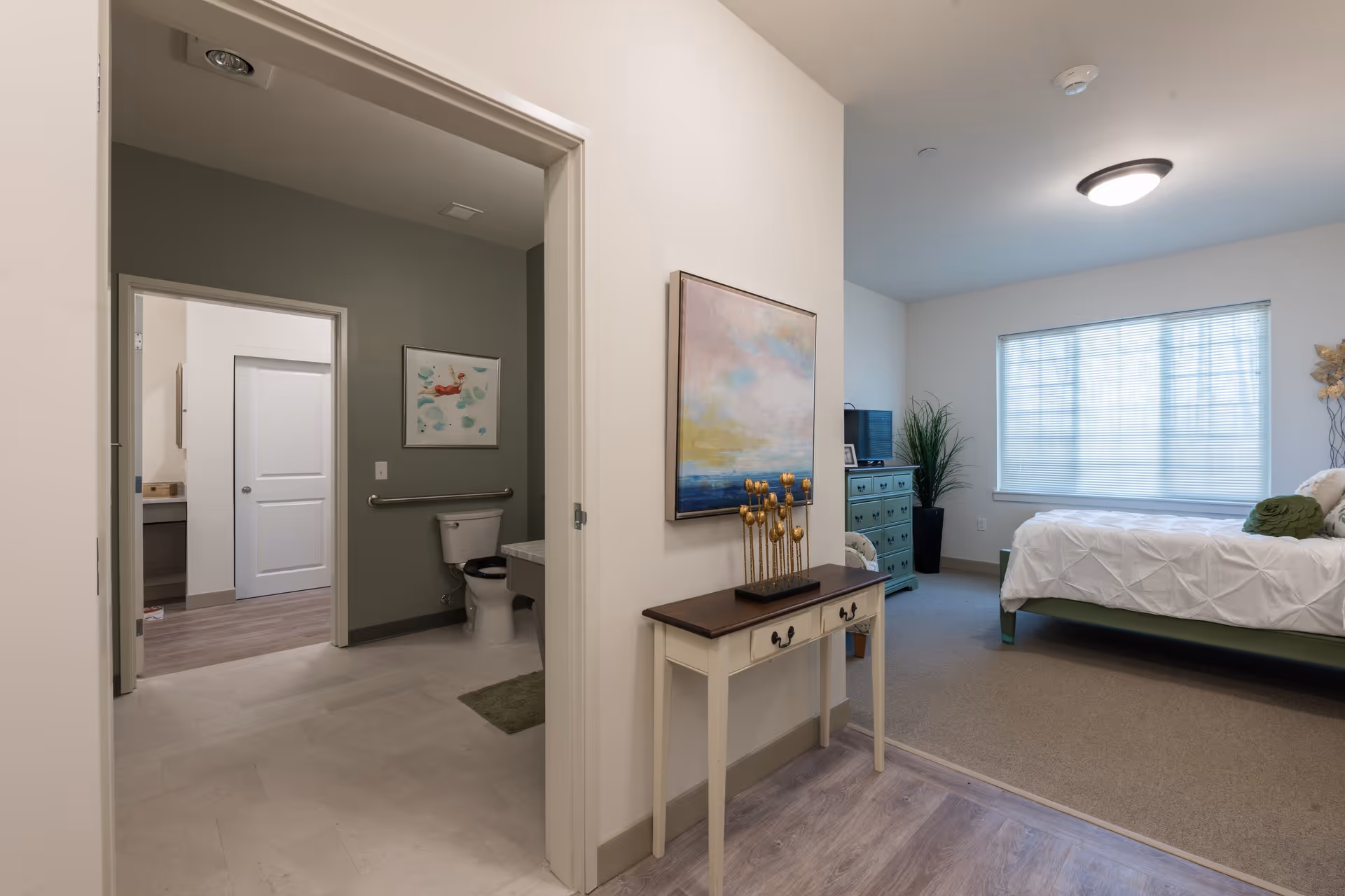 View of a senior living facility room showing a bedroom with a bed, dresser, TV, and a large window with blinds. Adjacent to the bedroom is a bathroom with a toilet, grab bar, and artwork on the wall. A small table with decorative items and a painting is positioned in the hallway between the bedroom and bathroom.