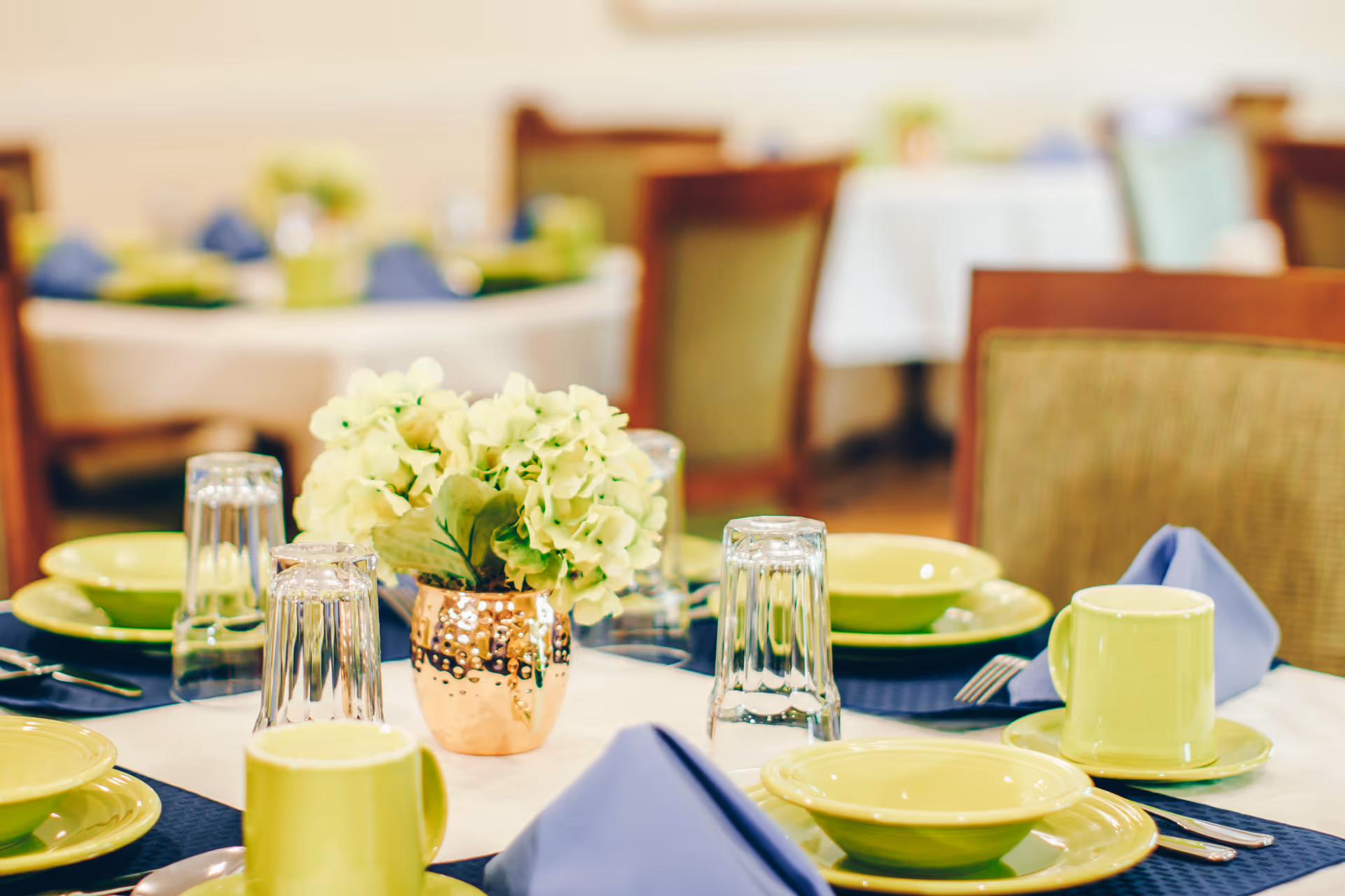 A dining table set with yellow plates, bowls, and mugs, blue cloth napkins, upside-down clear drinking glasses, silverware, and a small copper vase holding light green hydrangea flowers in the center. The background shows more tables and chairs in a softly lit dining room.