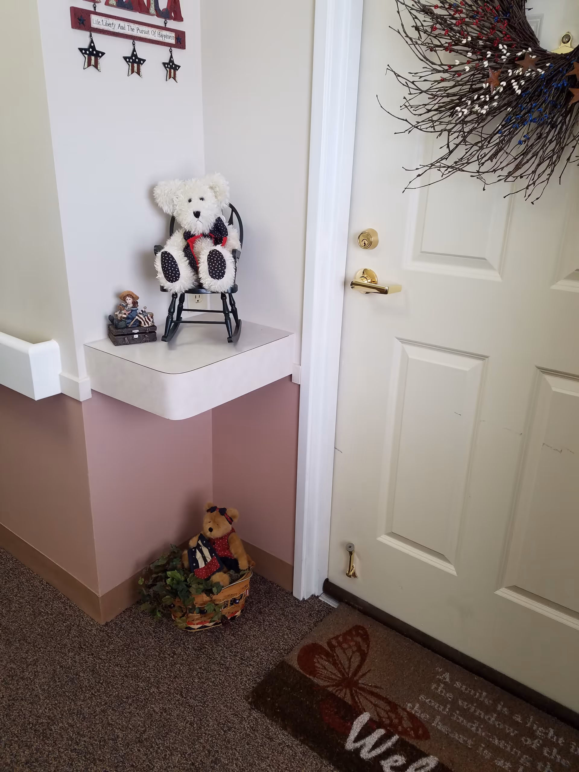 An entryway corner showing a white door with a wreath, a welcome mat, and decorative teddy bears on a shelf and in a basket.