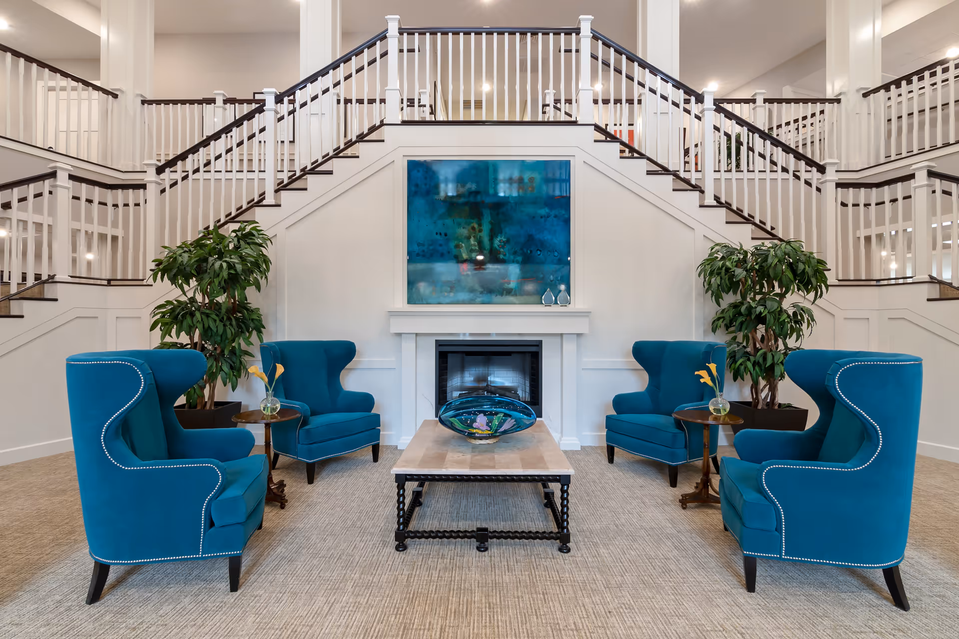 A spacious and elegant senior living facility common area featuring four teal upholstered armchairs arranged around a rectangular coffee table with a decorative glass centerpiece. Behind the seating area is a white fireplace with a large blue abstract painting above it. Two large potted plants flank the fireplace, and a grand staircase with white railings and dark wooden handrails ascends on both sides in the background.
