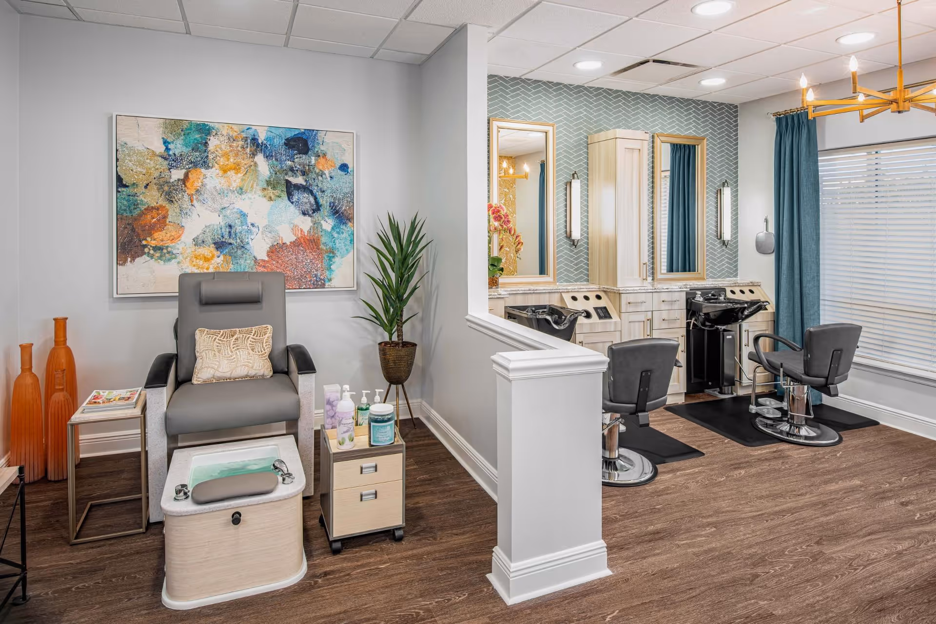 A modern salon area in a senior living facility featuring a pedicure chair with foot bath, a side table with lotions and supplies, two styling chairs in front of mirrors with sinks, decorative vases, a potted plant, and a large colorful abstract painting on the wall.