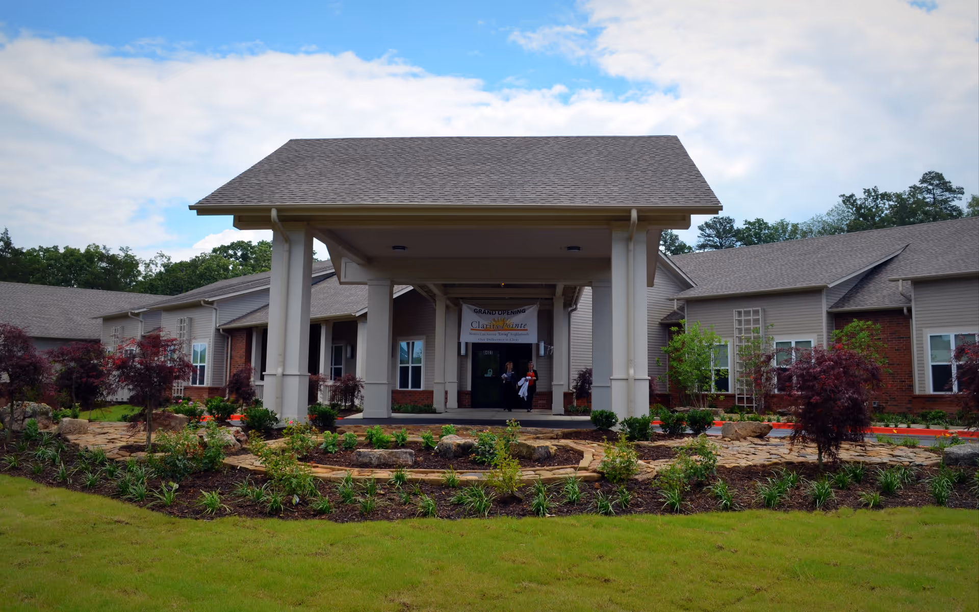 Front exterior view of a single-story senior living facility building with a covered entrance supported by columns. The building has a combination of brick and siding walls, multiple windows, and a landscaped garden with shrubs and small trees in front. Two people are visible near the entrance under a banner.