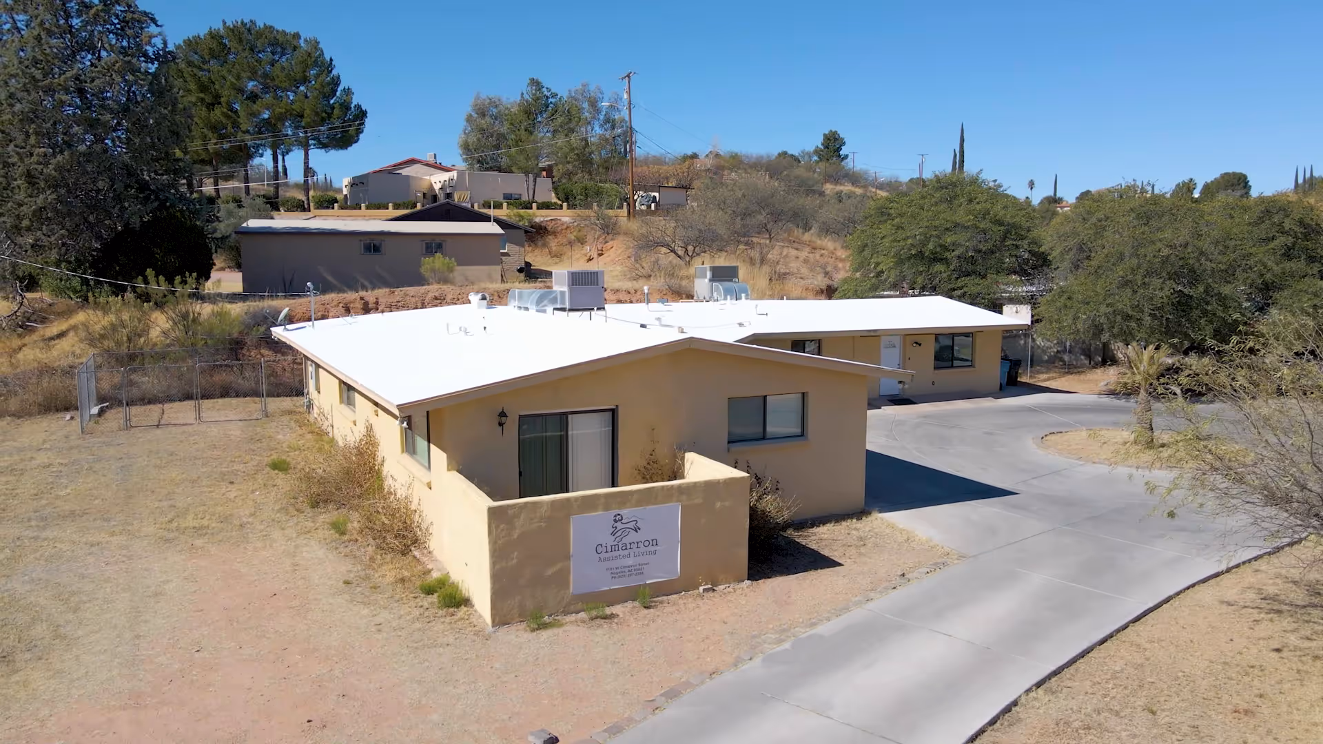 Exterior view of a single-story beige building with a white roof, surrounded by dry grass and trees under a clear blue sky. A sign on the front wall reads 'Cimarron Assisted Living'. A paved driveway curves around the building.