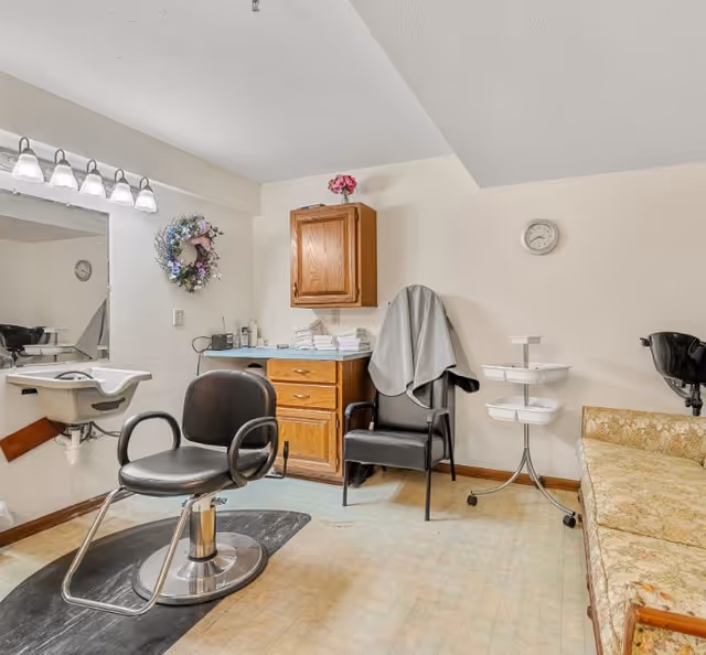 Small salon-style room with a barber chair in front of a sink and mirror, a wooden cabinet, additional seating, and styling supplies.