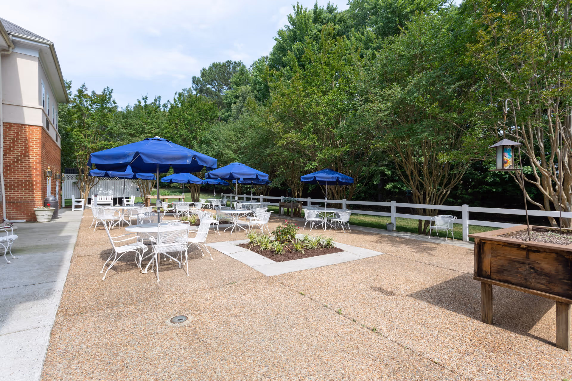 Outdoor patio with white metal tables and chairs, several blue umbrellas, planters, and trees along a fence by a building.