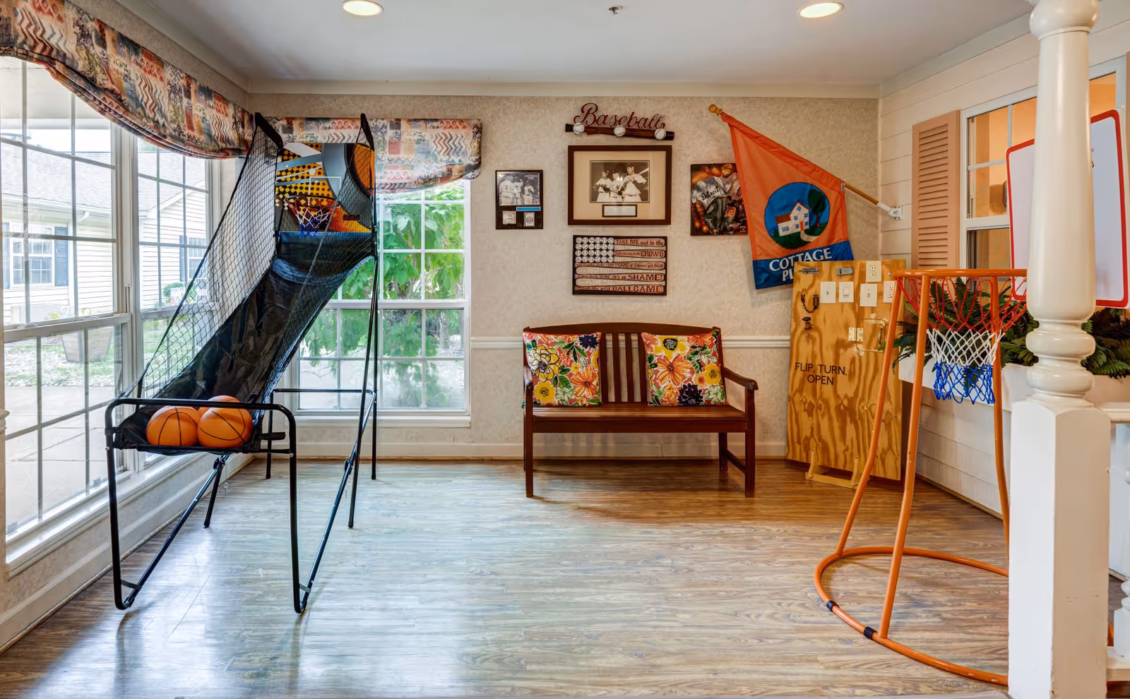 Indoor recreational area with a basketball arcade game holding three basketballs, a small orange basketball hoop, a wooden bench with two floral cushions, and various baseball-themed decorations on the wall including framed pictures and a flag. Large windows with patterned valances let in natural light.
