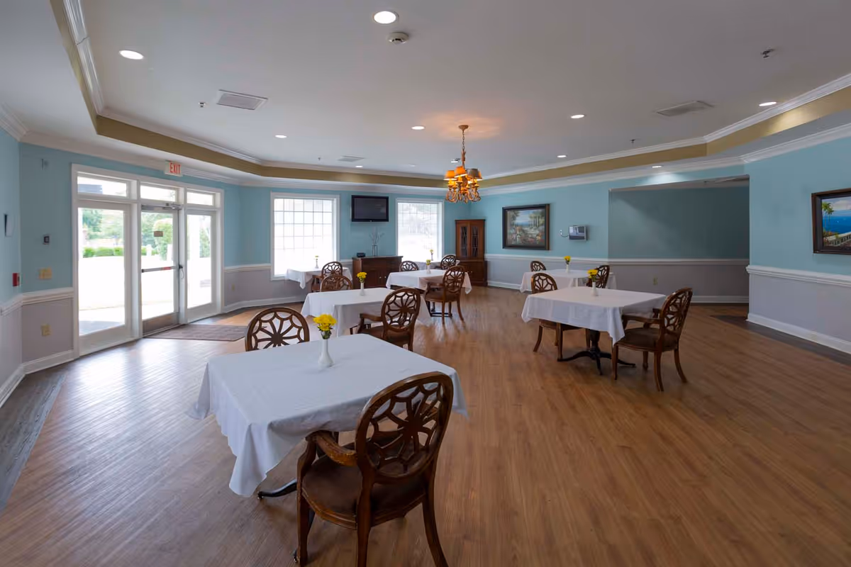A dining room with several tables covered in white tablecloths, each with a small vase of yellow flowers. The room has light blue walls with white trim, wooden chairs with decorative backs, wood flooring, a chandelier, and large windows letting in natural light. There is a TV mounted on the wall and framed paintings on the walls.