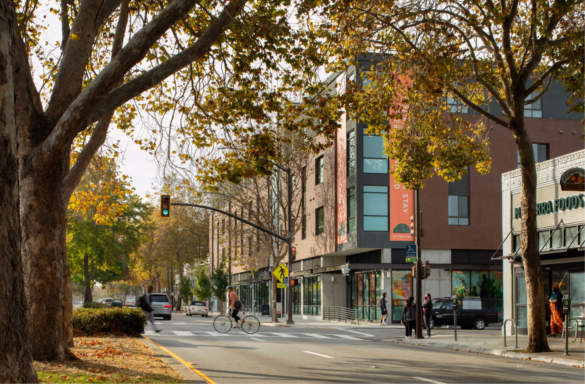 A street view showing a modern multi-story building with large windows and a red vertical banner on the corner. Several people are walking and a cyclist is crossing the street at a crosswalk. Trees with autumn leaves line the street, and a traffic light shows green. Nearby is a building with a sign reading 'Merrera Foods'.