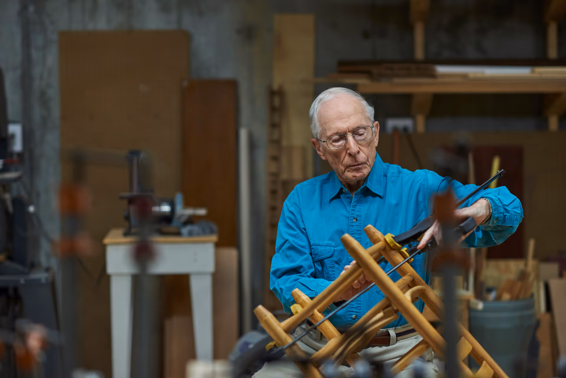 An elderly man wearing glasses and a blue shirt is working on a wooden chair in a workshop filled with woodworking tools and materials.