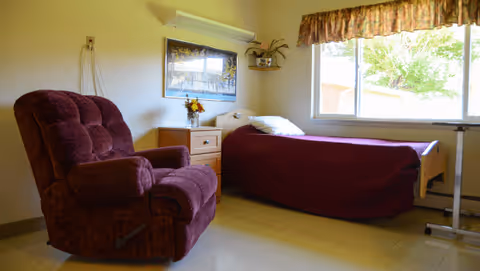 A nursing facility room with a single bed covered in a dark red blanket, a matching dark red recliner chair, a wooden nightstand with a vase of flowers, a framed picture on the wall, a small wall shelf with a plant, and a large window with floral curtains letting in natural light.