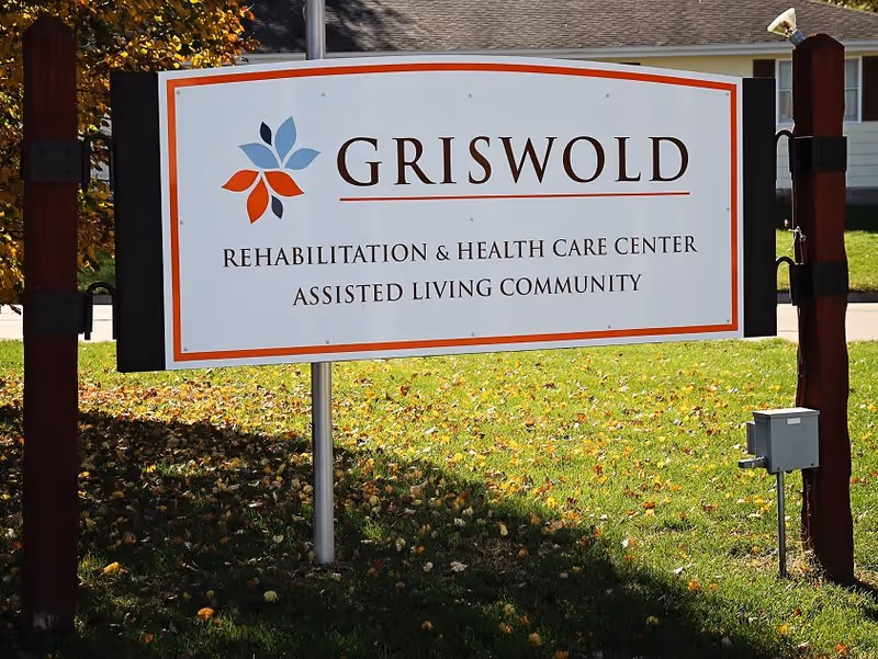 Outdoor sign for Griswold Rehabilitation & Health Care Center Assisted Living Community, mounted on wooden posts with grass and a building in the background.
