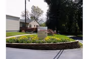 Outdoor view of a senior living facility named Mosser, showing a landscaped area with yellow flowers and a sign indicating parking and visitor parking. The building and trees are visible in the background under a clear sky.
