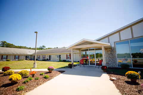 Exterior view of Heritage House of Portage Assisted Living facility on a sunny day, showing the entrance with a covered porch, landscaped flower beds with colorful flowers, and a clear blue sky.
