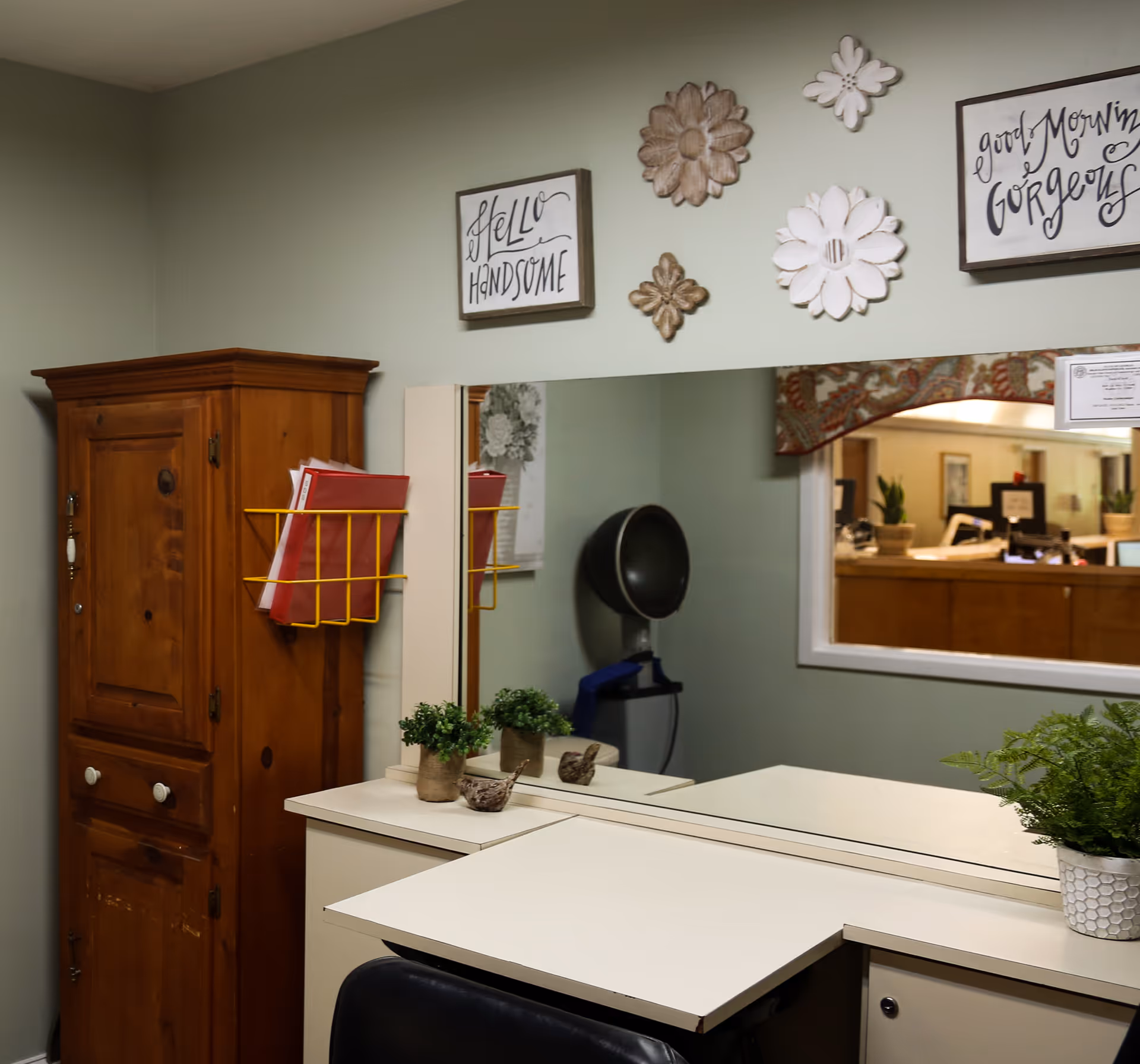 Interior view of a room with a wooden cabinet, a white desk with a mirror, decorative flower wall hangings, and framed signs that say 'Hello Handsome' and 'Good Morning Gorgeous'. There are small potted plants on the desk and a hair dryer visible in the reflection of the mirror.