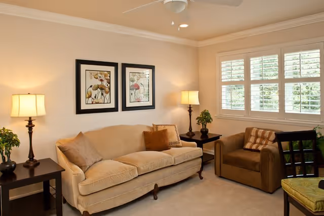 A cozy living room with a beige sofa and a brown armchair, both adorned with decorative pillows. Two framed floral artworks hang on the wall above the sofa. There are two side tables with lamps and small plants on either side of the sofa. A window with white shutters lets in natural light, and a ceiling fan is visible above.