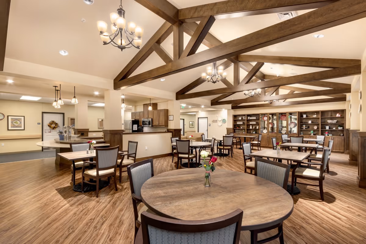 A spacious dining room with wooden floors and exposed wooden ceiling beams. Several round and square tables with chairs are arranged throughout the room, each table decorated with a small vase of flowers. The room features warm lighting from chandeliers and pendant lights. In the background, there is a kitchen area with cabinets and appliances, and a large wooden cabinet displaying plates and decorative items.