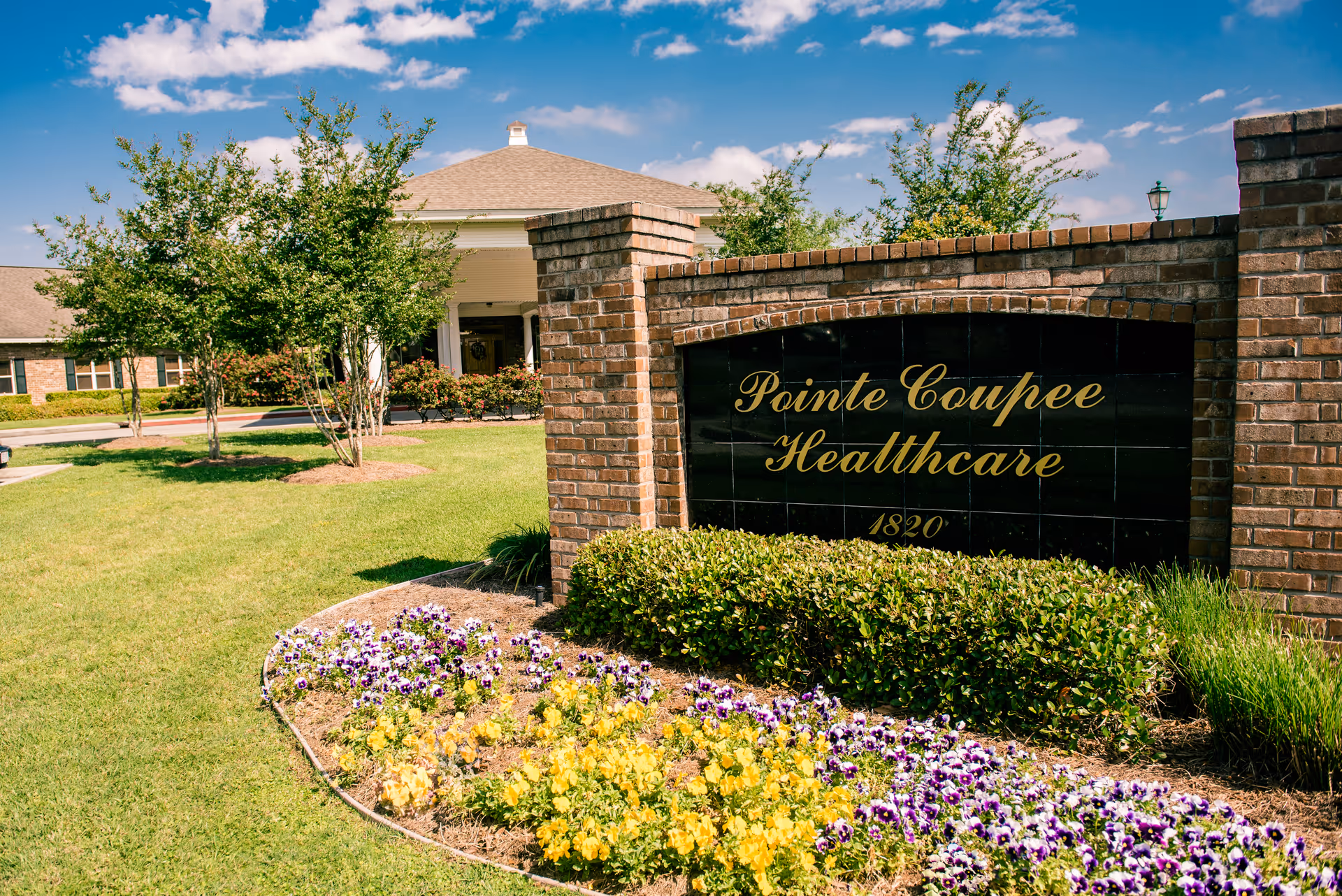 Brick entrance sign reading 'Pointe Coupee Healthcare' with landscaped flowers and the facility building in the background.