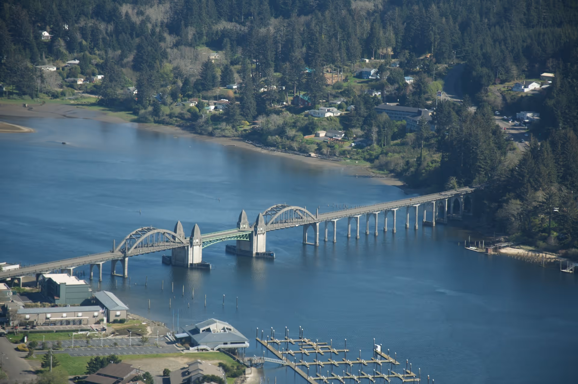 Aerial view of a long bridge spanning across a wide river with a marina and buildings on one side and a forested area with houses on the other side.