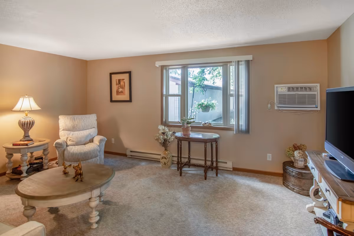A cozy living room with beige walls and carpeted floor. The room features a comfortable armchair next to a round side table with a lamp and books. In front of the armchair is an oval wooden coffee table with small decorative figurines. A window with vertical blinds lets in natural light, and beneath it is a small wooden table with potted plants. On the right side, there is a wall-mounted air conditioner above a round wicker basket and a flat-screen TV on a wooden TV stand.