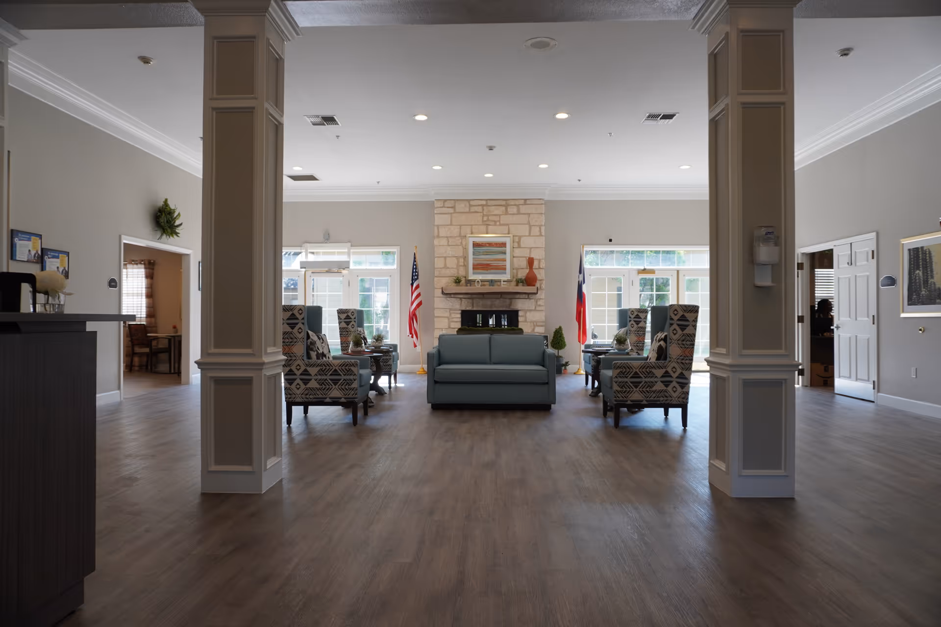 A spacious and well-lit common area in a senior living facility with a central stone fireplace, a blue sofa, and patterned armchairs arranged around small tables. The room has wooden flooring, two large windows with glass doors on either side of the fireplace, and American and Texas flags placed near the windows. There are two white columns in the foreground and neutral-colored walls with some framed artwork and decorations.