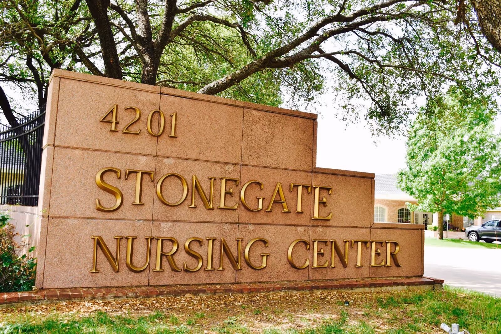 A large stone sign with gold lettering that reads '4201 Stonegate Nursing Center' situated outdoors with trees and a building visible in the background.