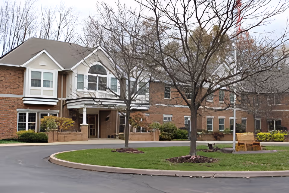 Exterior view of Sprenger Health Care Village of The Falls showing a two-story brick building with white trim, a covered entrance, leafless trees, a circular driveway, and a green lawn with a wooden bench.