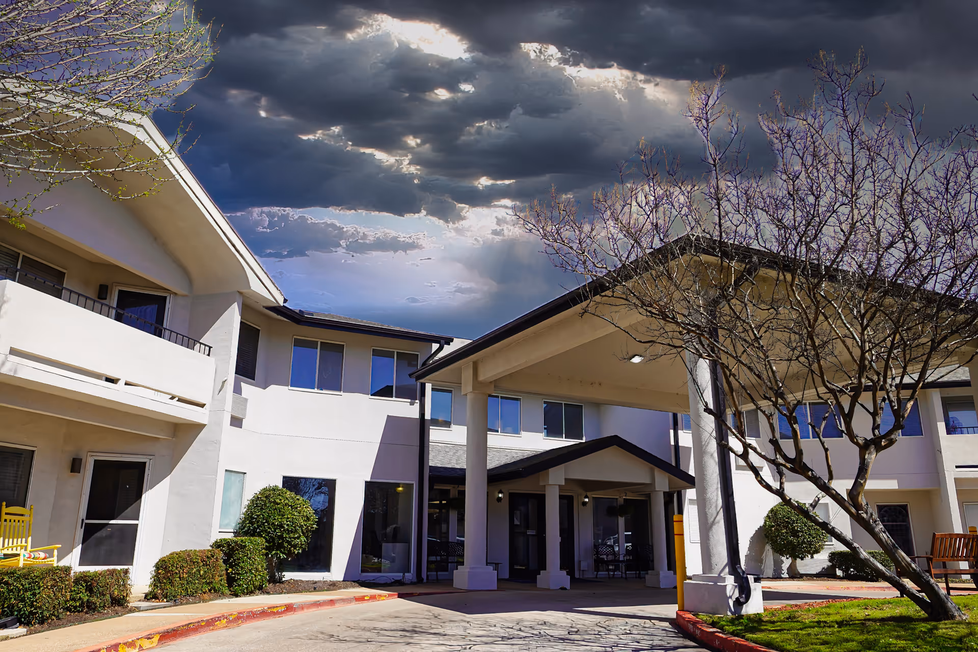 Exterior view of a two-story senior living facility building with white walls, large windows, and a covered entrance. There are trimmed bushes and a leafless tree in the landscaped area near the entrance. The sky above is partly cloudy with rays of sunlight breaking through.