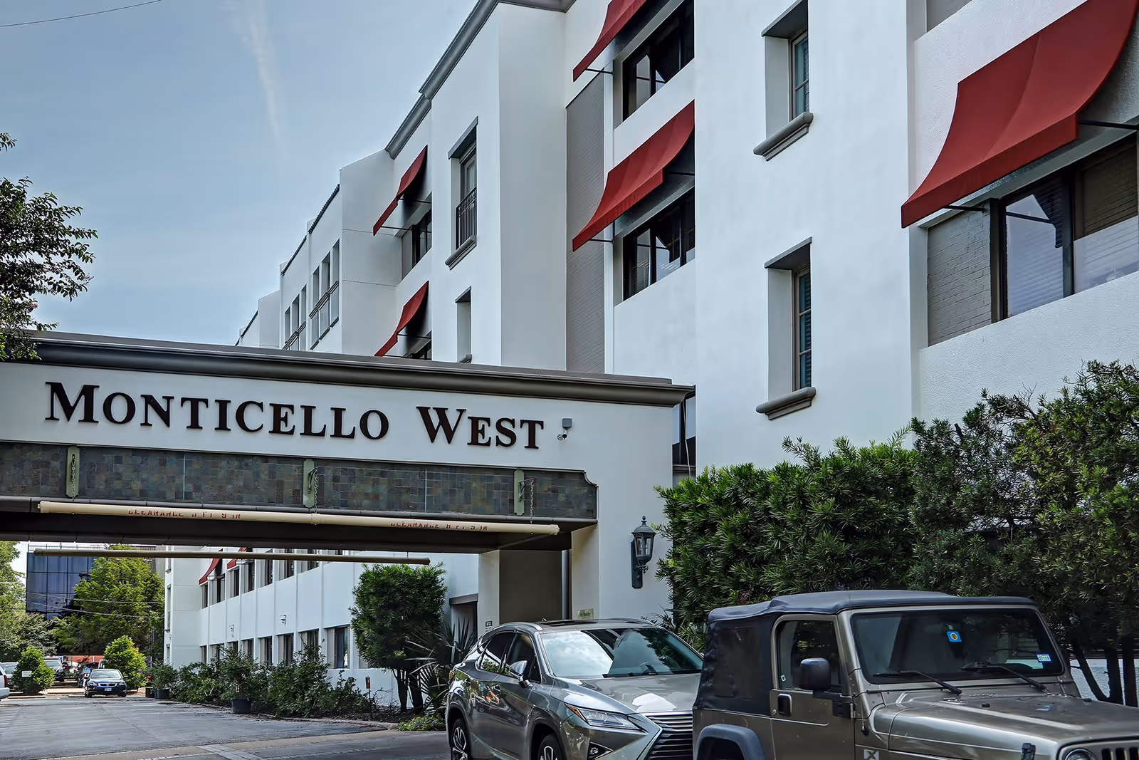 Exterior view of Monticello West building with white walls and red window awnings, cars parked in front, and greenery along the side.