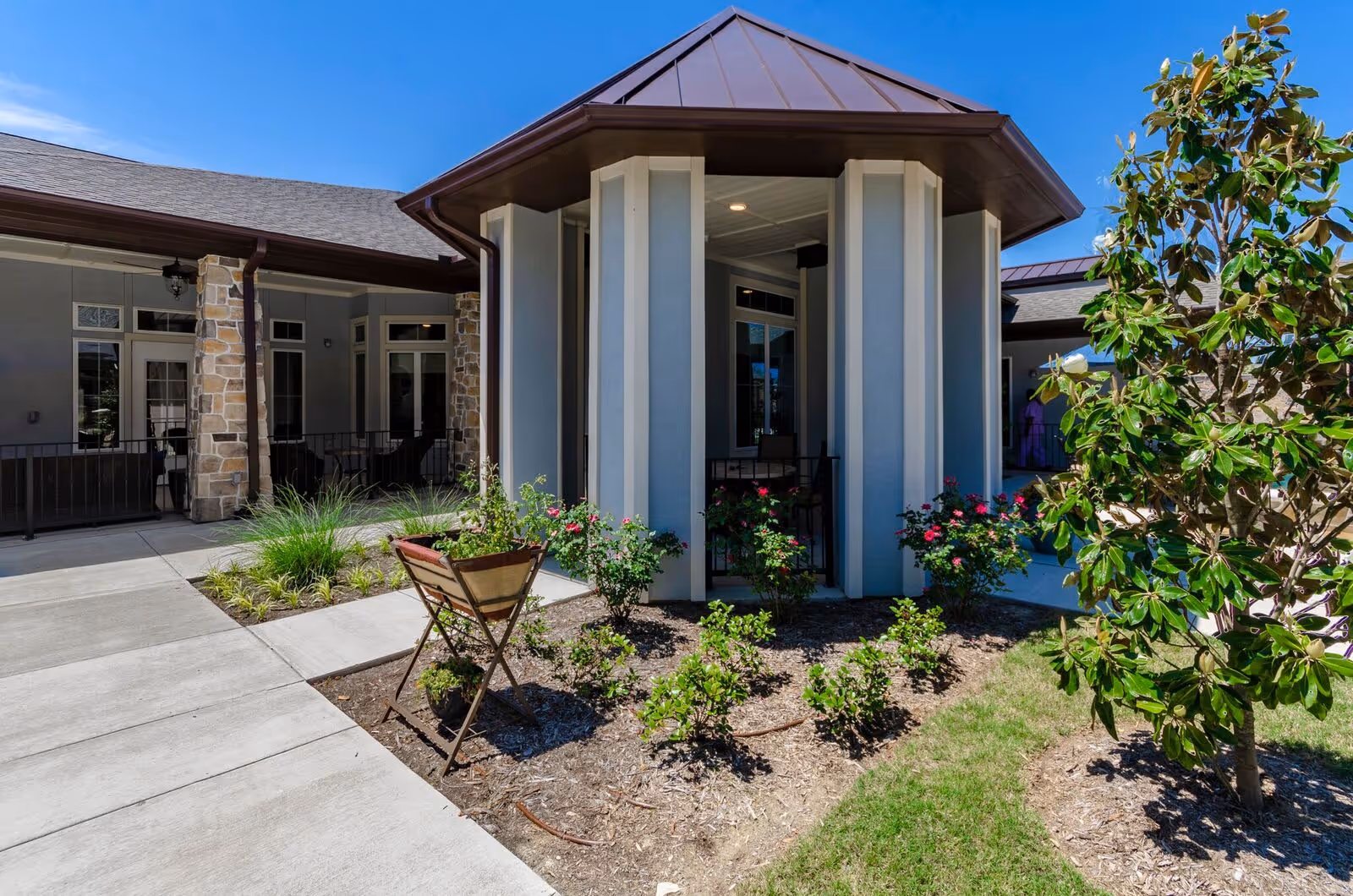 Outdoor courtyard area of a senior living facility with a small garden featuring flowering plants and a tree. The building has stone pillars, large windows, and a covered walkway under a clear blue sky.