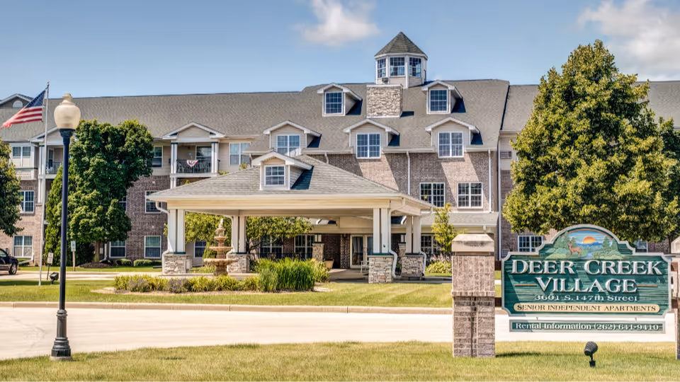 Front exterior of a multi-story assisted living building with a covered porte-cochere, fountain, and a 'Deer Creek Village' sign on the lawn.