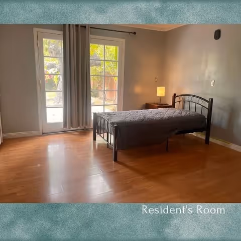 A simple resident's bedroom with a single bed covered in a gray quilt, a wooden nightstand with a lit lamp, and large glass doors with curtains leading outside. The room has wooden flooring and gray walls.