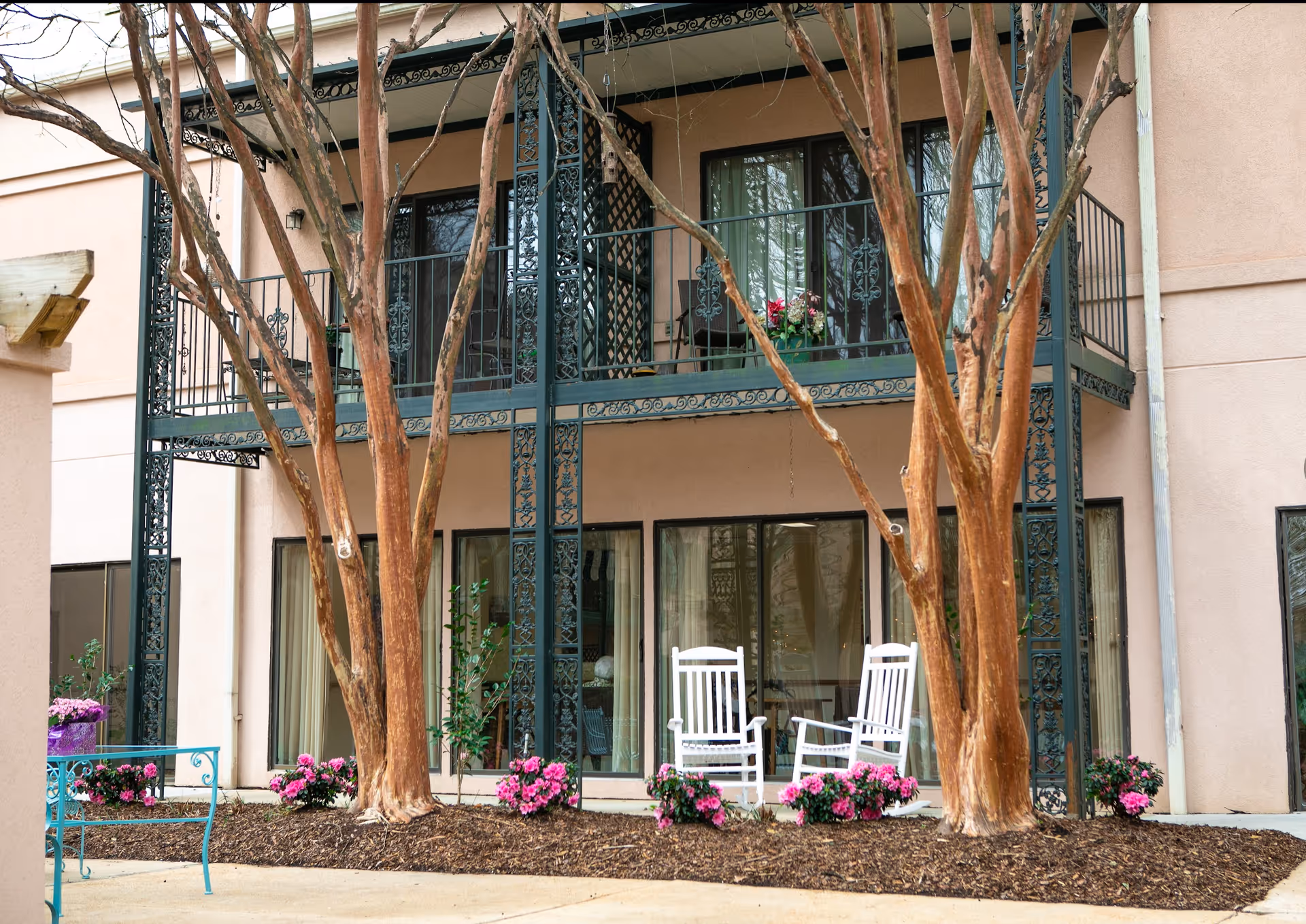 Two white rocking chairs and potted flowers on a ground-floor patio beneath an ornate iron balcony with trees in front of a beige building.