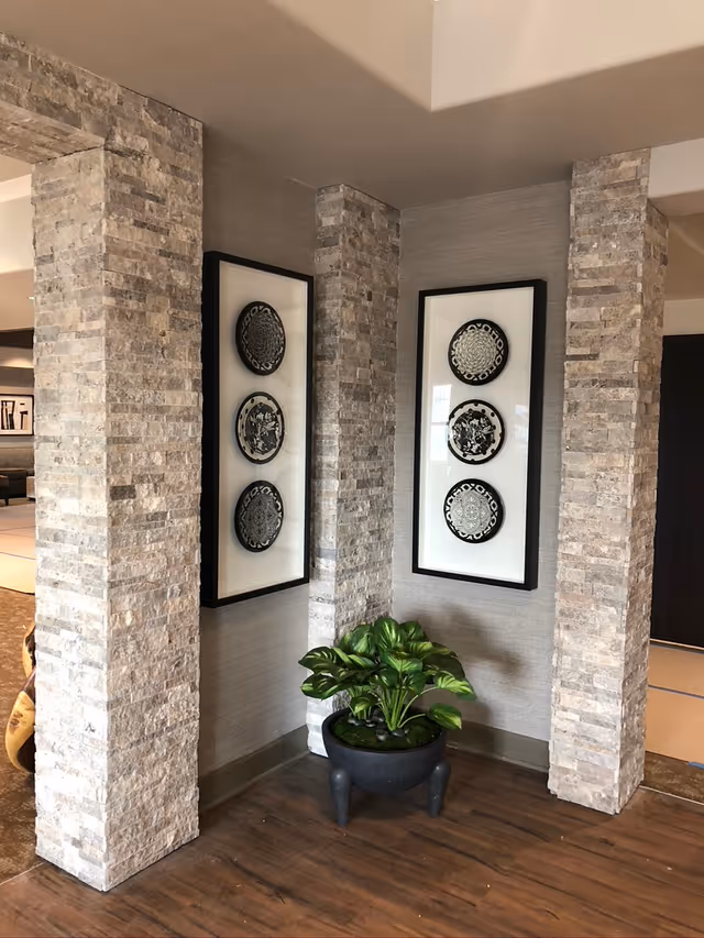 Corner of an interior space with three stone pillars and two framed wall decorations featuring three circular black and white patterned plates each. A potted green plant sits on the wooden floor beneath the wall art.