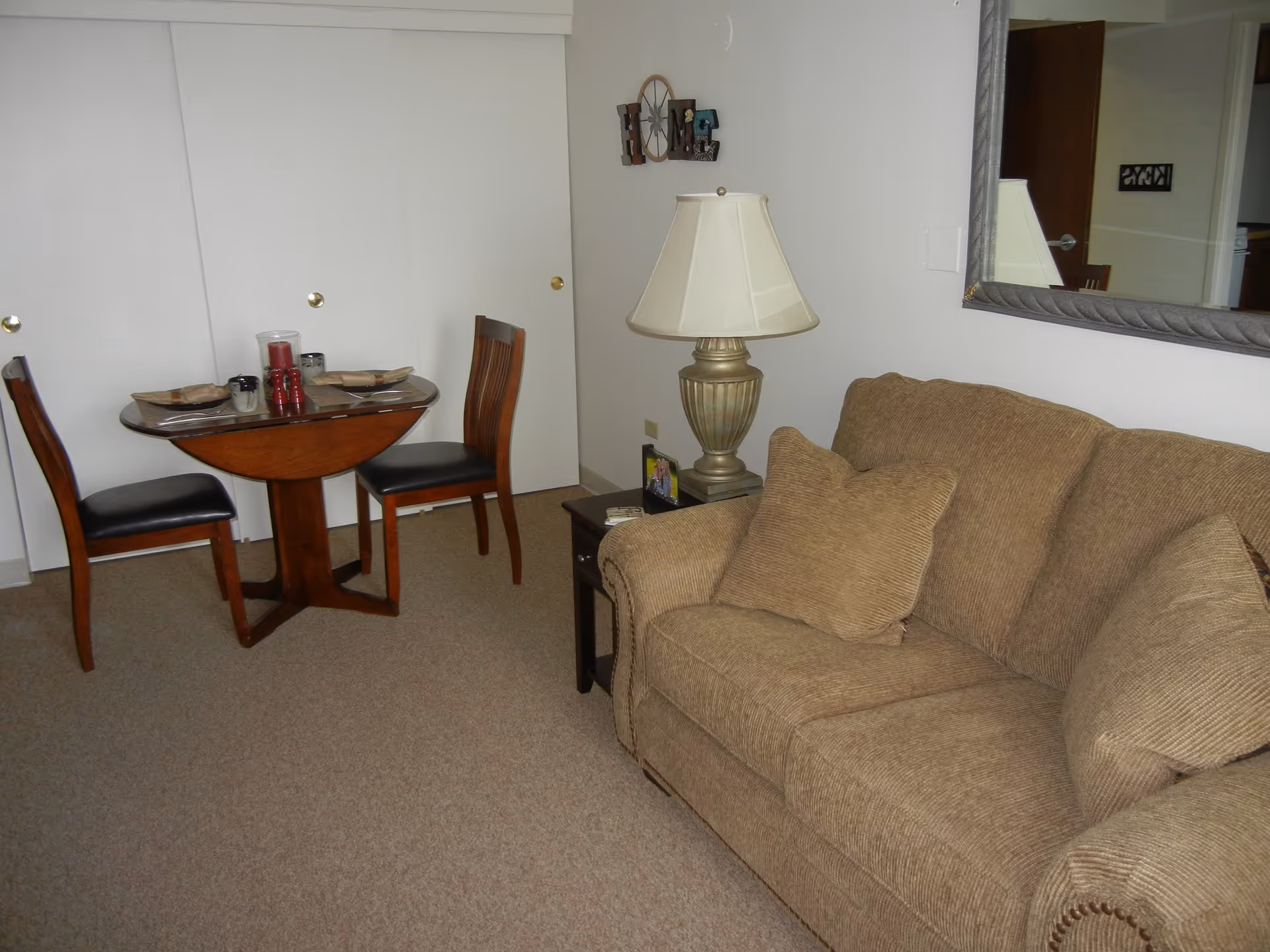 A cozy living area with a brown upholstered couch with two matching pillows, a side table with a lamp and a framed photo, and a small round wooden dining table set for two with chairs. A large mirror hangs on the wall above the couch, and a decorative 'HOME' sign is mounted on the adjacent wall.