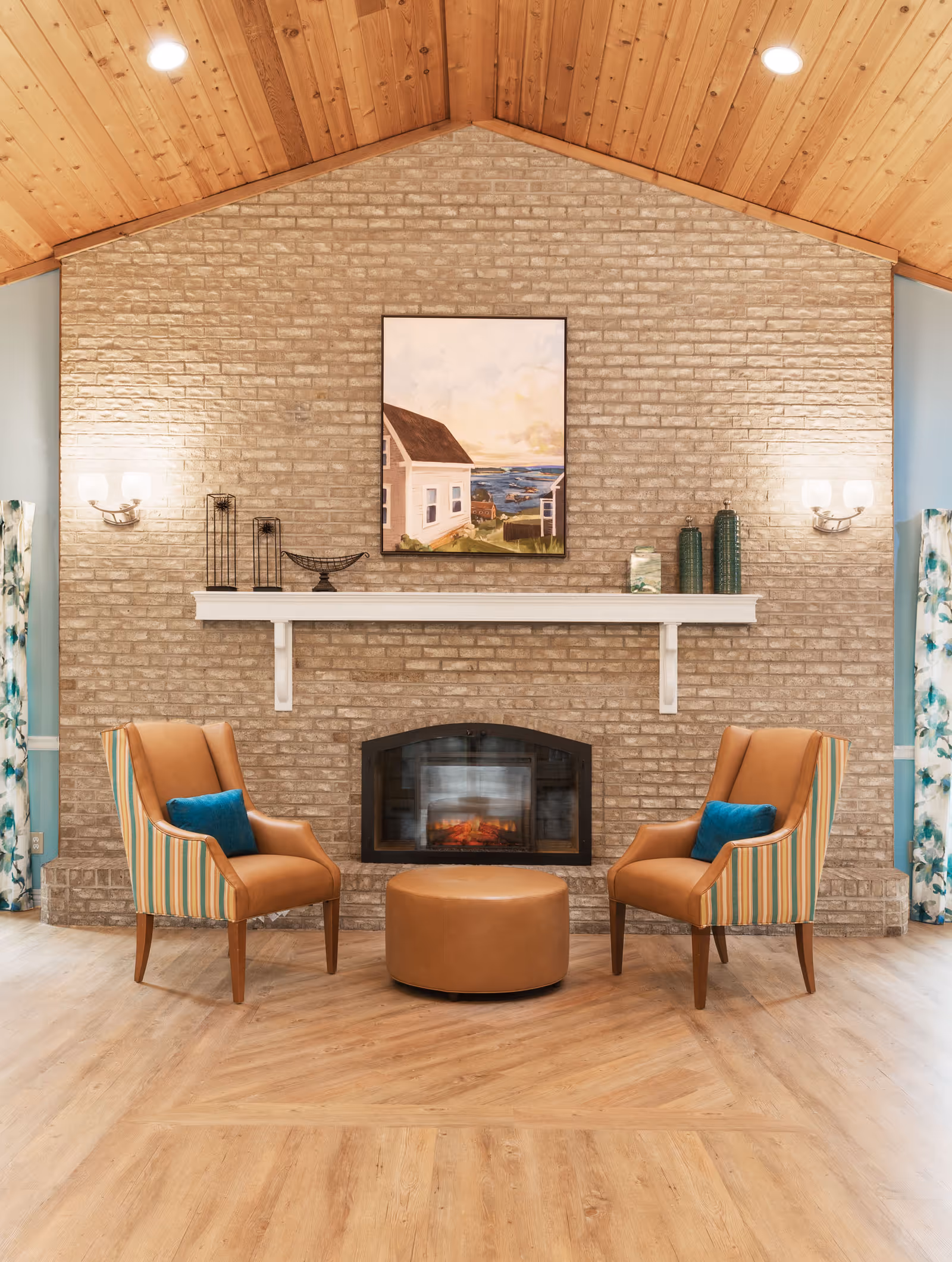 A cozy seating area with two striped armchairs featuring blue cushions facing each other across a round brown ottoman. Behind them is a brick fireplace with a white mantel, decorated with vases and a decorative bowl. Above the mantel hangs a painting of a house by the sea. The room has a wooden vaulted ceiling with recessed lighting and light wood flooring.