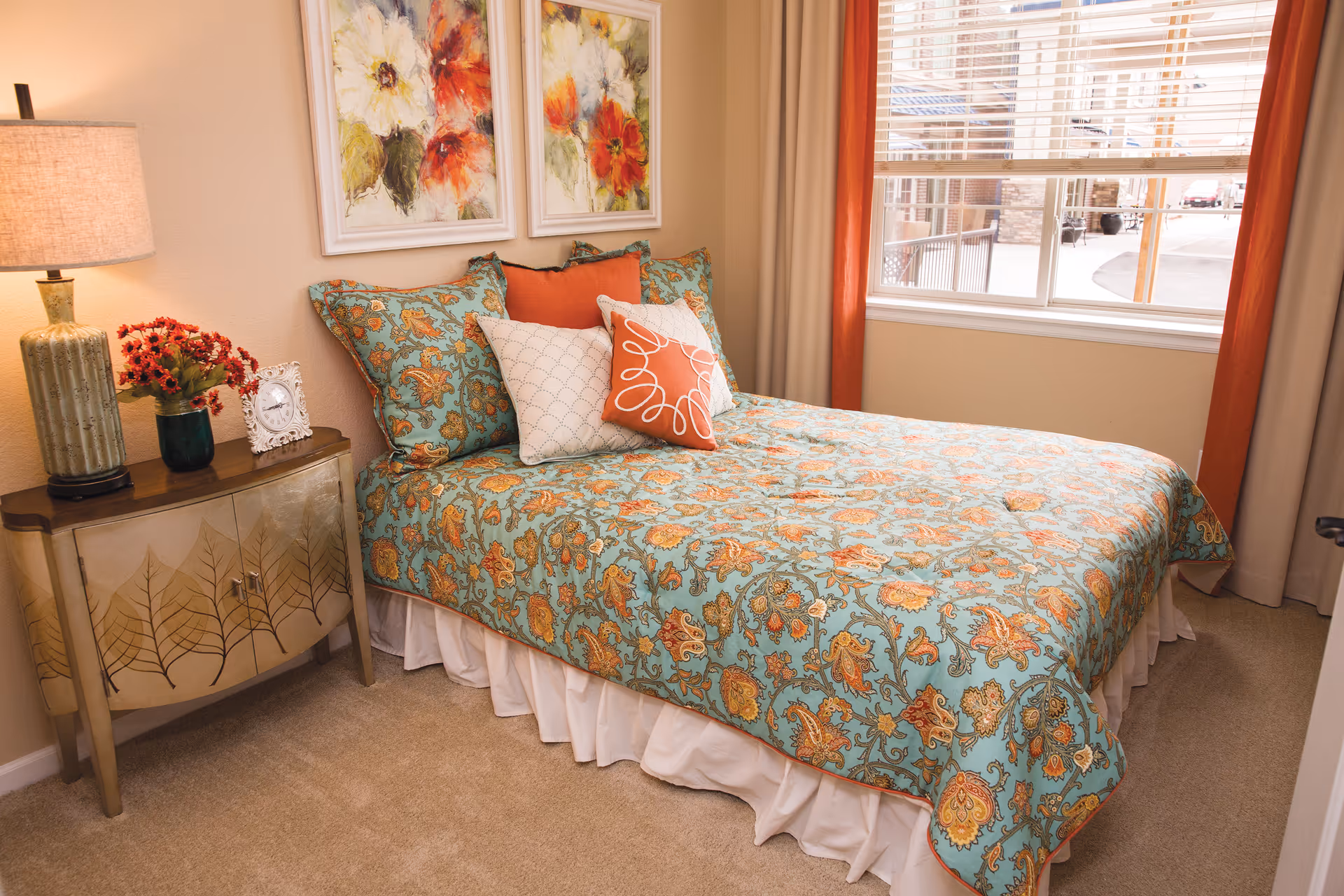 A cozy bedroom with a bed covered in a blue and orange floral patterned bedspread, accented with matching pillows. Next to the bed is a wooden nightstand with a lamp, a small vase of red flowers, and a decorative clock. Two floral paintings hang on the wall above the bed. A window with beige and orange curtains lets in natural light.