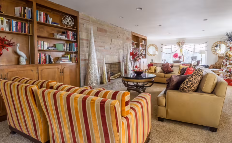 A cozy living room with a striped sofa and a beige armchair adorned with leopard print and brown pillows. The room features a stone fireplace with decorative tall cone-shaped ornaments, built-in wooden bookshelves filled with books and decor, a round black coffee table, and large windows letting in natural light. The space is decorated with festive garlands and flowers, creating a warm and inviting atmosphere.