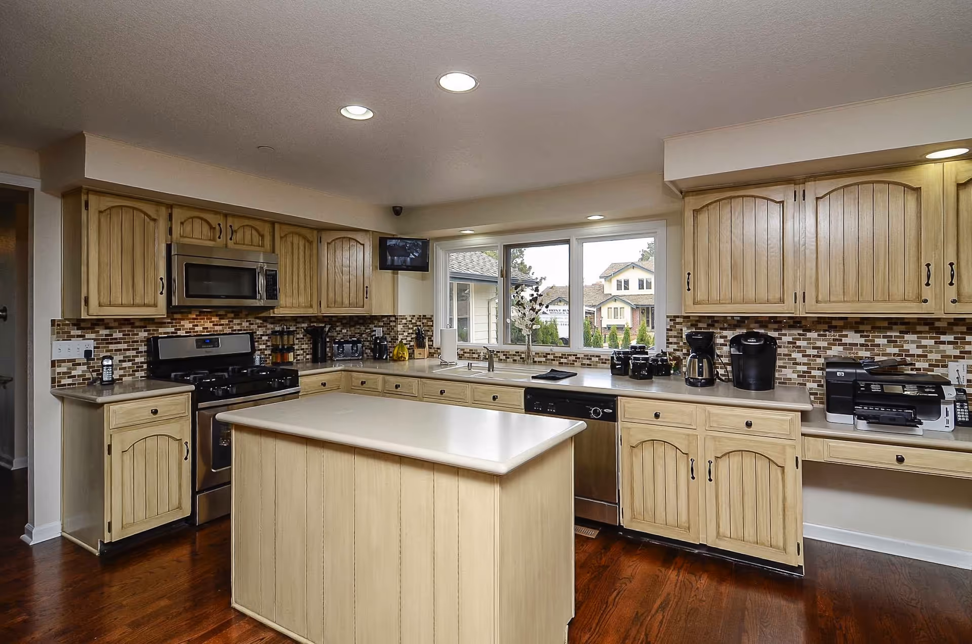 A spacious kitchen with light wood cabinets, a central island, stainless steel appliances including a stove and dishwasher, a coffee maker, and a printer. The kitchen has a mosaic tile backsplash and a large window overlooking houses outside. The floor is wooden and the ceiling has recessed lighting.