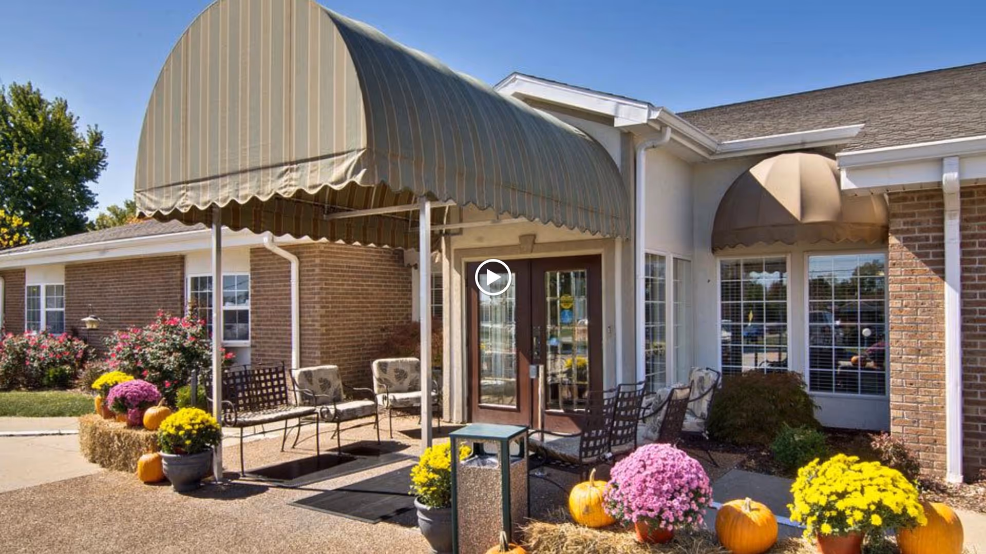 Outdoor entrance area of a senior living facility with a striped canopy over the door, metal chairs with cushions arranged outside, potted yellow and purple flowers, pumpkins, and hay bales decorating the walkway. The building has brick and white siding with windows and a smaller rounded canopy over one window.