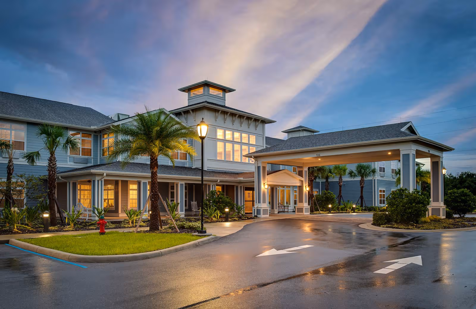Exterior view of Sancerre at Orange City senior living facility at dusk, showing a large, well-lit building with multiple windows, palm trees, a covered entrance, and a driveway with directional arrows.