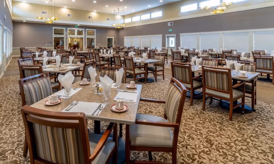 Spacious dining room with multiple tables set for meals, each table having cups, plates, silverware, and folded napkins. The room has patterned carpet, large windows with blinds, and modern ceiling lights.