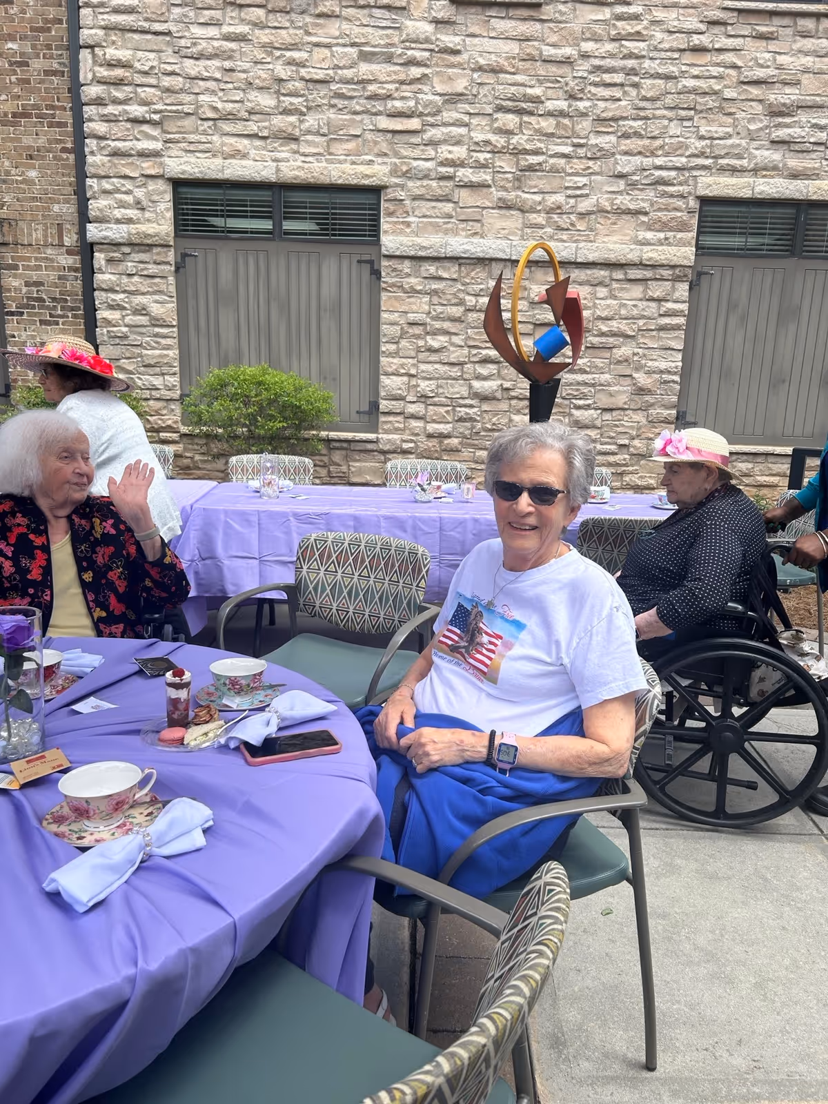 Several elderly women sitting around tables covered with purple tablecloths outdoors near a stone building wall. One woman in sunglasses and a white t-shirt with an American flag design is smiling at the camera. Another woman in a wheelchair wears a pink hat, and a third woman waves while seated. The tables have teacups, napkins, and small desserts on them.