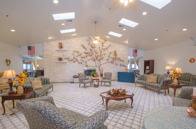 A spacious and well-lit living room area in a senior living facility with patterned carpet, multiple upholstered sofas and chairs arranged around wooden coffee tables, decorative lamps, and floral arrangements. The back wall features a large stone facade with a metal tree wall art and small shelves with decorative items. Skylights and ceiling fans are visible on the high ceiling.